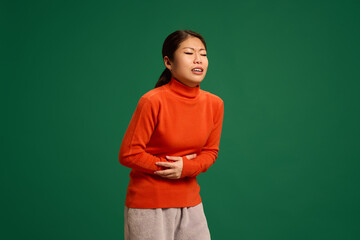 Young, pretty Asian woman clutching stomach, grimacing in pain, illustrating stomach discomfort or...