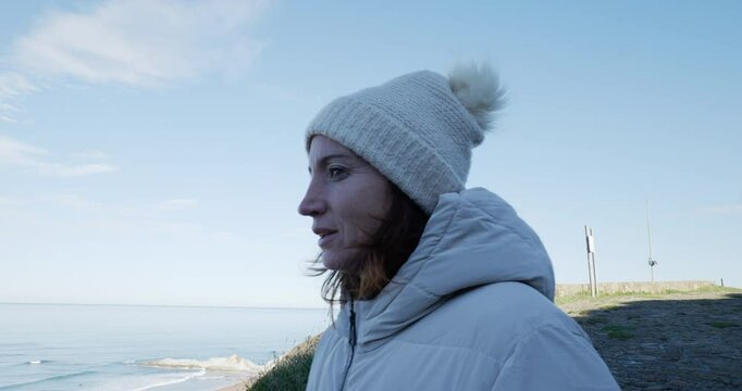 Mujer con gorro y chaqueta de abrigo desde la sombra mira al mar en los primeros d&iacute;as de primavera en un d&iacute;a soleado, plano de perfil 