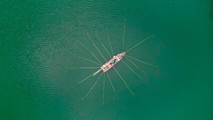 A fisherman balances on a small boat in the middle of a tranquil lake, managing multiple fishing rods as the calm waters reflect the serene surroundings.