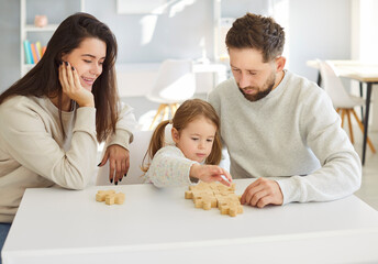 Portrait of a little cute child girl collecting wooden jigsaw with mother and father sitting at...
