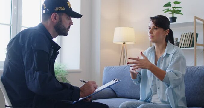 Very emotional upset shocked young woman witness talks to police officer who is investigating crime case and taking notes while interviewing her. Red blue lights flashing over living room