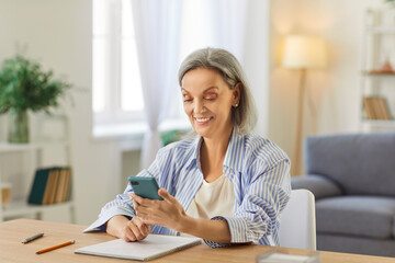 Portrait of happy smiling senior woman in casual clothes sitting at the desk in the living room at...