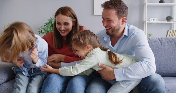 Happy family of four having fun together. Parents and children spending time at home. Cheerful little girl tickles her brother while sitting on the couch with mother and father. Family love concept