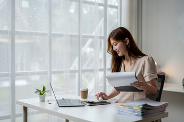 A woman is sitting at a desk with a laptop and a stack of papers