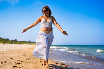 Summertime by sea. Beautiful middle-aged woman walking on sandy beach on sunny summer day. Front...