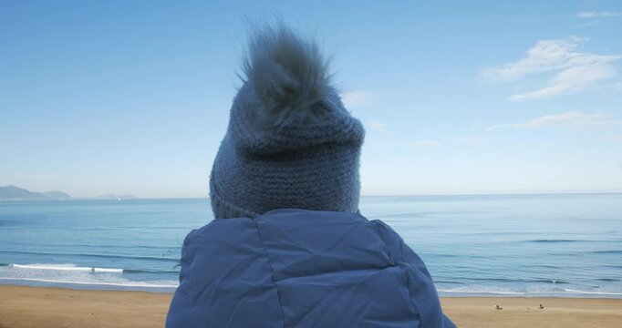 Mujer con gorro y chaqueta de abrigo desde la sombra mira al mar en los primeros d&iacute;as de primavera en un d&iacute;a soleado, plano desde atr&aacute;s 