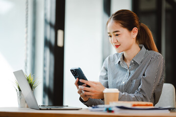Obraz premium Cheerful beautiful Asian woman uses smartphone to chat at her office. Female accountant makes annual financial and tax reports.