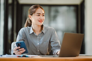 Cheerful beautiful Asian woman uses smartphone to chat at her office. Female accountant makes annual financial and tax reports.