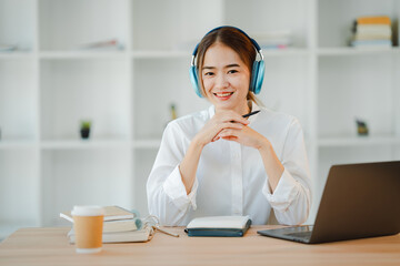 Naklejka premium Beautiful Asian female student with a cheerful smile Wearing headphones, studying online and taking notes at the library. Female accountant makes annual financial and tax reports.