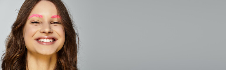 A young woman smiles with unique pink eyebrows against a grey background, banner