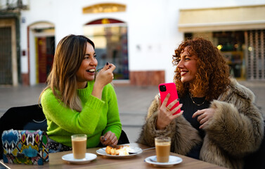 two girls sitting on the terrace of a bar while drinking coffee and cake while looking at each other smiling