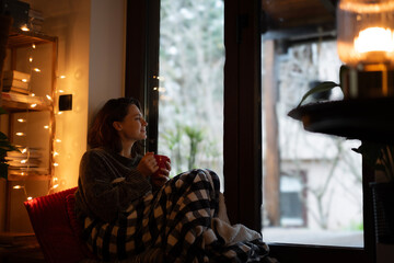 Thoughtful woman drinking a cup of coffee at home while sitting near window, countryside lifestyle