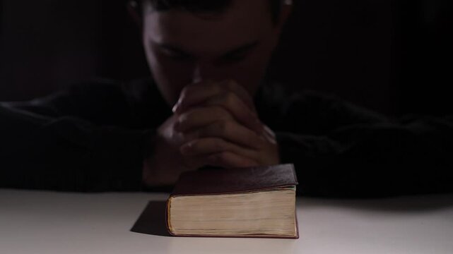 Young man praying with holy bible on white table in dark room