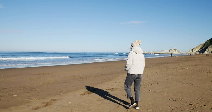 Mujer feliz con gorro y chaqueta de abrigo llega a la playa al en los primeros d&iacute;as de primavera en un d&iacute;a soleado, y se quita el gorro y la chaqueta, frente al mar. Plano general.