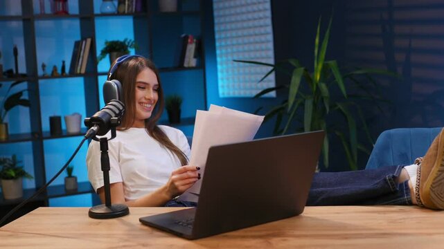 Relaxed, holding paper with text. Woman is hosting radio podcast with microphone and laptop.