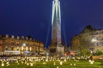 Night scene of a tall war memorial, illuminated and surrounded by small lights in a town square, Beam Light Festival, Harrogate, Yorkshire, UK.