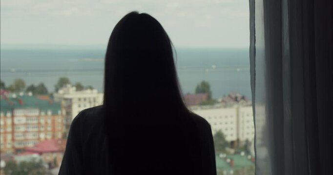 Woman opening curtains and looking at cityscape by the sea