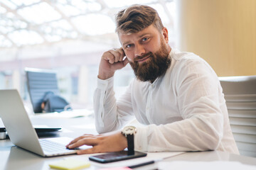 Fototapeta premium Portrait of handsome male employee posing at table desktop with digital netbook computer on it,Caucasian man in smart casual apparel looking at camera using coworking internet connection for freelance
