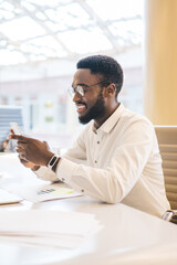 Happy dark skinned male in glasses for vision protection checking mobility during remote job on laptop technology in modern coworking space, cheerful man enjoying smartphone messaging indoors