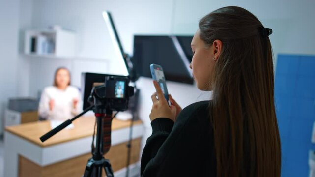 Brunette girl takes video on her phone of the display of professional camera. Female blogger speaks to camera at blurred backdrop.