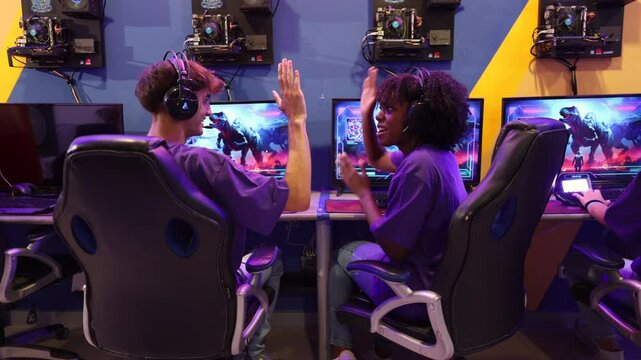 Two excited gamers wearing headphones are shaking hands and celebrating victory after winning an esports competition in an internet cafe, surrounded by other players