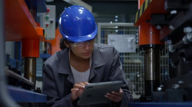 Indian woman engineer with a blue helmet holding a tablet while checking and controlling working machine parts and tools, close up shot.