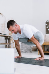 Strong caucasian male in casual wear spending free time on yoga training watching online lessons on laptop at home interior, pensive man 20s concentrated on practice holding body on hands in asana