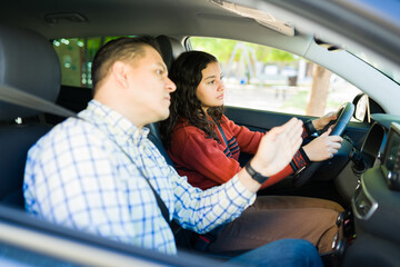Latin father teaching his teenage daughter how to drive inside their car, the girl is holding the...