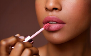 A close-up of a woman applying lipstick. She has a well-defined, full set of lips painted in a soft pink shade. The woman is using a pink-colored lipstick applicator to apply the lipstick.