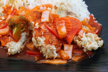 Oriental dish: spaony chicken, vegetables, sweet and sour sauce, salad and rice on a plate stands on the table.