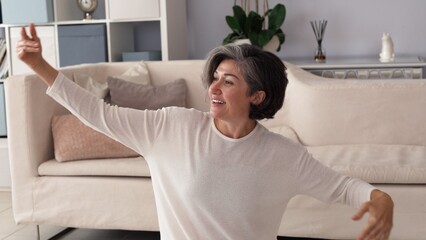 Caucasian woman, gray streaked hair, white sweater, arms gracefully lifted, smiling softly in...