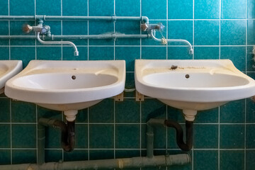 Two white sinks in an outdated bathroom with green tiles showing wear and plumbing details