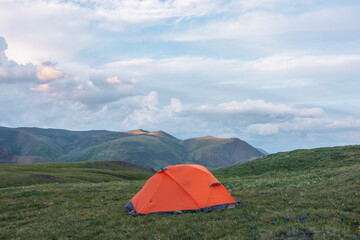Vivid orange tent on grassy flowering hill with view to high multicolor mountain under cloudy sky. Sunlit mountain tops of sunset colors in changeable weather. Twilight landscape with colorful tops.