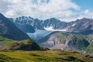 Colorful landscape with orange tent in alpine valley with view to big glacier tongue and large snow-capped mountain range under clouds in blue sky. Green hills and rocks against ice and sheer crags.