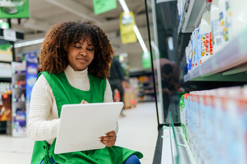 Beautiful black woman working in a supermarket. Female assistant using a digital tablet for taking inventory of products in display rack in a grocery store.