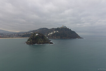 Fototapeta premium Scenic view of Santa Clara Island and Monte Igueldo with turquoise waters and coastal cityscape under a cloudy sky in San Sebastian, Spain.