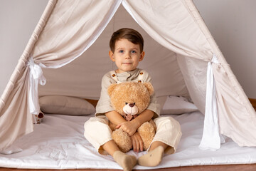 Happy 5 years old little boy sitting in a wigwam tent, hugging soft toy teddy bear. Children's...