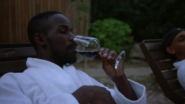 Homosexual LGBT male couple enjoying a resort weekend, relaxing after a hot tub treatment, tasting and drinking red wine on a terrace, handheld shot.