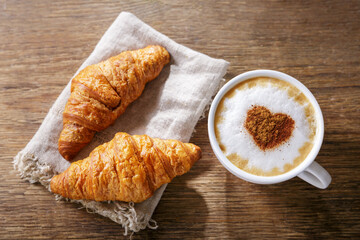 Cup of cappuccino coffee with drawn heart and fresh croissants, top view