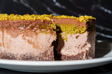 Slice of chocolate Dubai cheesecake with pistachio paste and kataifi dough filling covered in chocolate ganache on white plate. Close up. Indulgence, gourmet desserts, art of culinary presentation