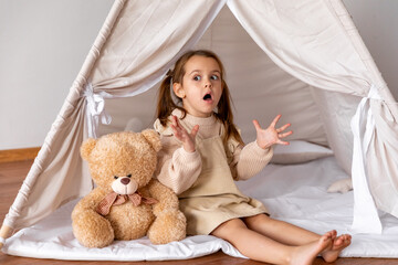 Happy surprised 4-5 years old little girl sitting in a wigwam tent, near soft toy teddy bear....