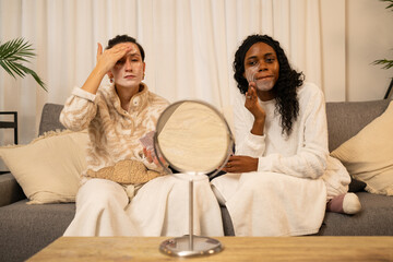 Diverse female friends sharing skincare moment, applying moisturizing face cream while relaxing together in comfortable home setting, bonding through wellness ritual