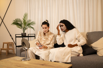 Obraz premium Female friends lounging together, wearing bathrobes and applying facial masks during skincare routine in cozy living room setting