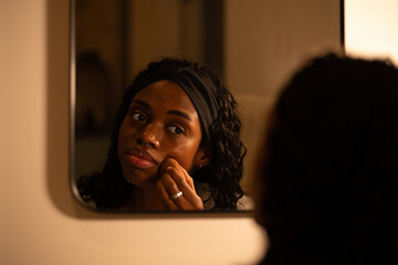 Young woman with curly hair and a headband examining her skin while touching her face in the bathroom mirror at night, focusing on her skincare routine and imperfections