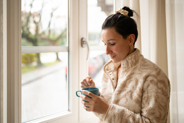 Comfortable fleece clad woman stirring steaming mug, gazing through window at rain soaked street...