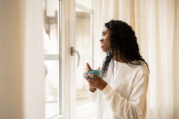 Young woman with curly hair drinking coffee, sitting near sunlit window, experiencing peaceful...