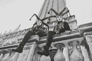 Black and white photo of two masked figures in elaborate jester costumes sitting on marble...