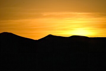 Silhouettes of sand dunes gently rolling across the horizon, illuminated by the warm glow of a golden sunset. The serene desert landscape reflects the quiet beauty of nature’s tranquility.