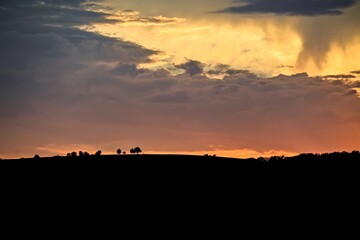 Dramatic evening sky with layers of dark clouds, illuminated by warm orange and yellow hues. A line of silhouetted trees atop a distant hill adds depth to this striking natural scene.