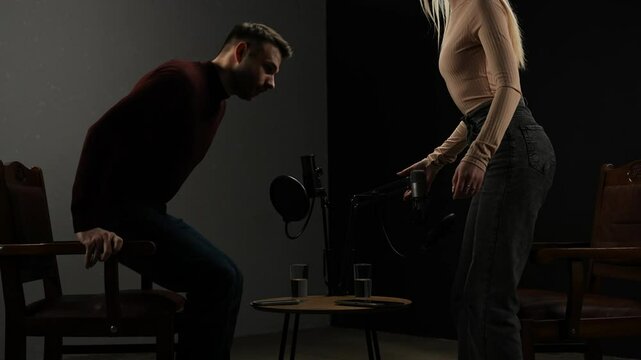 Man host and woman guest recording interview in stylish podcast studio using microphones, creating digital content, fixing mics and have conversation before start of the show.